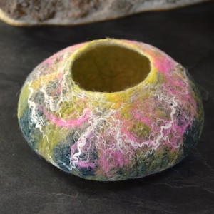 May include: A round, felted bowl with a light green interior and a colourful exterior. The bowl is decorated with swirls of pink, white, and green wool.