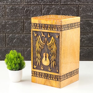 May include: A wooden box with a carved design of a guitar with wings and musical notes. The box is made of light brown wood and has a natural finish. The text "GLOBAL WOOD CRAFT" is engraved on the top of the box.