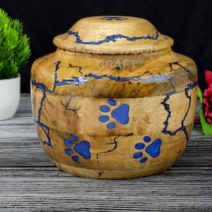 May include: Wooden pet urn with a blue paw print design. The urn has a lid and is made of natural wood with blue resin accents.