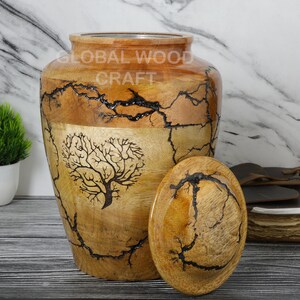 May include: A wooden urn with a tree design and a lid. The urn is made of wood with a natural finish and has a dark brown tree design on the front. The lid is also made of wood and has a natural finish.