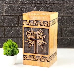 Könnte beinhalten: Eine Urne aus Holz mit einem Kreuz- und Blumendesign. Die Urne ist aus Naturholz gefertigt und hat eine dunkelbraune Beize. Der Text "GLOBAL WOOD CRAFT" ist auf der Oberseite der Urne eingraviert.