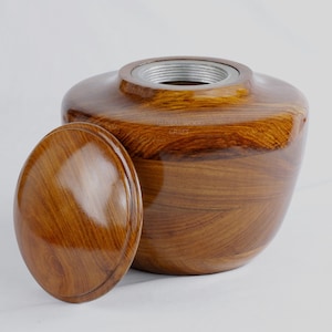 Puede incluir: Una urna de madera con tapa, que presenta una rica veta de madera marrón y una tapa de metal plateado. La urna está hecha a mano y tiene un acabado liso y pulido.