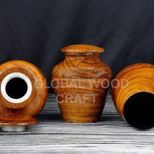 Puede incluir: Tres urnas de madera con tapas, cada una con un patrón único de veta de madera. Las urnas son marrones y tienen un acabado natural. El texto "GLOBAL WOOD CRAFT" es visible en la urna del medio.