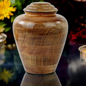 Può includere: Un'urna in legno con una finitura in legno naturale. L'urna è appoggiata su una superficie scura con due candele accese sullo sfondo.