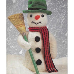 May include: A crocheted snowman wearing a green hat and a red scarf, with black buttons. It holds a broom with a green handle. The snowman has a carrot nose and a stitched smile. The background is a snowy scene.