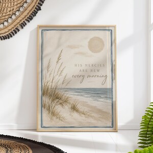 May include: A rectangular wall art print in a light wood frame featuring a coastal beach scene with tall tan beach grass, a blue ocean, and a pale sun. The text "HIS MERCIES ARE NEW every morning" is printed in the centre in dark brown lettering.