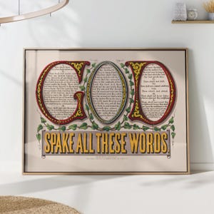 May include: Framed vintage print with the word "GOD" in large, ornate letters. The print features text from the Bible, with the phrase "SPAKE ALL THESE WORDS" at the bottom. The artwork is framed in a light-colored wooden frame.