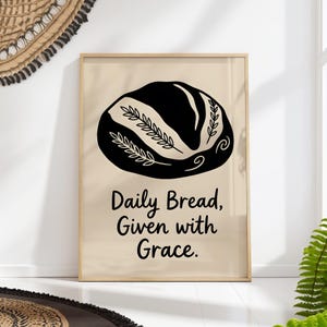 Puede incluir: Una lámina decorativa rectangular en un marco de madera clara con una ilustración gráfica negra de una hogaza de pan con detalles de espigas de trigo. Debajo del pan, el texto dice: Daily Bread, Given with Grace. Fondo de color beige.