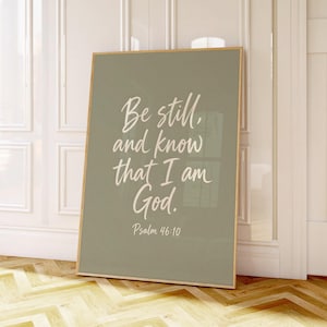 Puede incluir: Un cuadro decorativo rectangular con un marco de madera fino, fondo verde salvia y texto en cursiva blanca que dice: Be still, and know that I am God. Psalm 46:10. El diseño está centrado en la lámina con una disposición sencilla.