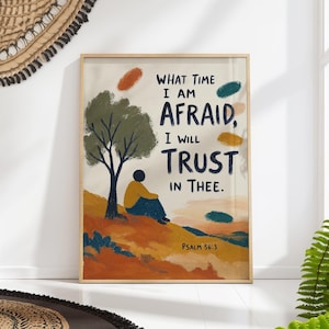 May include: A framed wall art print featuring a person sitting under a tree on a hill. The text reads: WHAT TIME I AM AFRAID, I WILL TRUST IN THEE. PSALM 56:3. The illustration uses a palette of olive green, mustard yellow, orange, and dark blue.