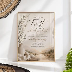 Op de afbeelding: Een ingelijste print met de tekst "Vertrouw op de Heer met heel je hart" uit Spreuken 3:5. Het kunstwerk toont een aquarel landschap met een vaas, bladeren en een rivierlandschap in zachte, neutrale tinten. De lijst is licht van kleur.