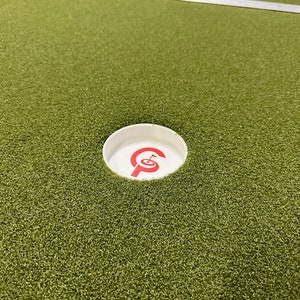 May include: A white golf hole with a red "G" logo on a green putting green.