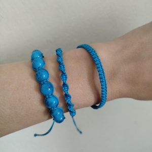 Peut inclure: Trois bracelets bleus sont présentés sur un poignet. Les bracelets comprennent un style perlé, un design en cordon torsadé et une bande tissée. Les perles sont d'un bleu turquoise vif, et les deux autres bracelets sont d'un bleu assorti.