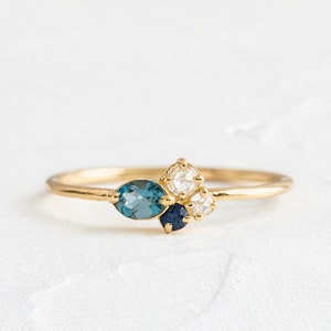 Può includere: Un anello d'oro con un gruppo di pietre preziose blu e bianche. L'anello presenta una pietra preziosa blu di forma ovale più grande, una pietra preziosa blu rotonda più piccola e due pietre preziose bianche rotonde più piccole.