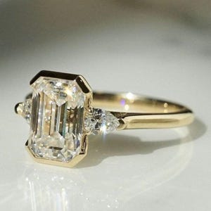 Puede incluir: Un anillo de compromiso de oro con un diamante de talla esmeralda y dos diamantes de acento en forma de pera.