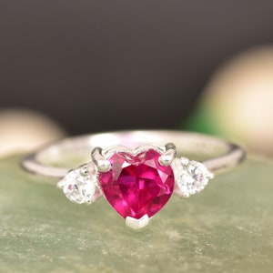 Puede incluir: Un anillo de plata con una piedra preciosa de rubí en forma de corazón y dos piedras blancas más pequeñas a cada lado.