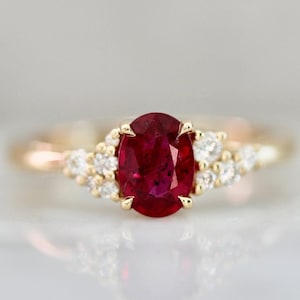 Op de afbeelding: Een gouden ring met een grote ovale robijn-edelsteen omringd door kleinere diamanten. De ring heeft een delicate, gedraaide band.