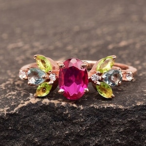 May include: A rose gold ring featuring a large oval ruby gemstone, surrounded by smaller green and blue gemstones.