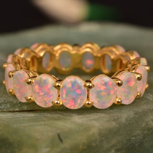 May include: A gold band ring with a row of oval-shaped opal gemstones. The opals are a pale pink color with iridescent flashes of color.