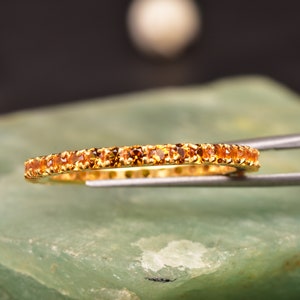 Puede incluir: Un anillo de banda de oro con una fila de pequeñas piedras preciosas de color naranja.