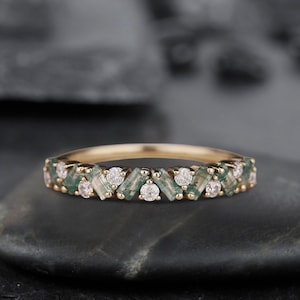 May include: A gold band ring with a row of alternating green and white gemstones. The ring is set on a gray background.
