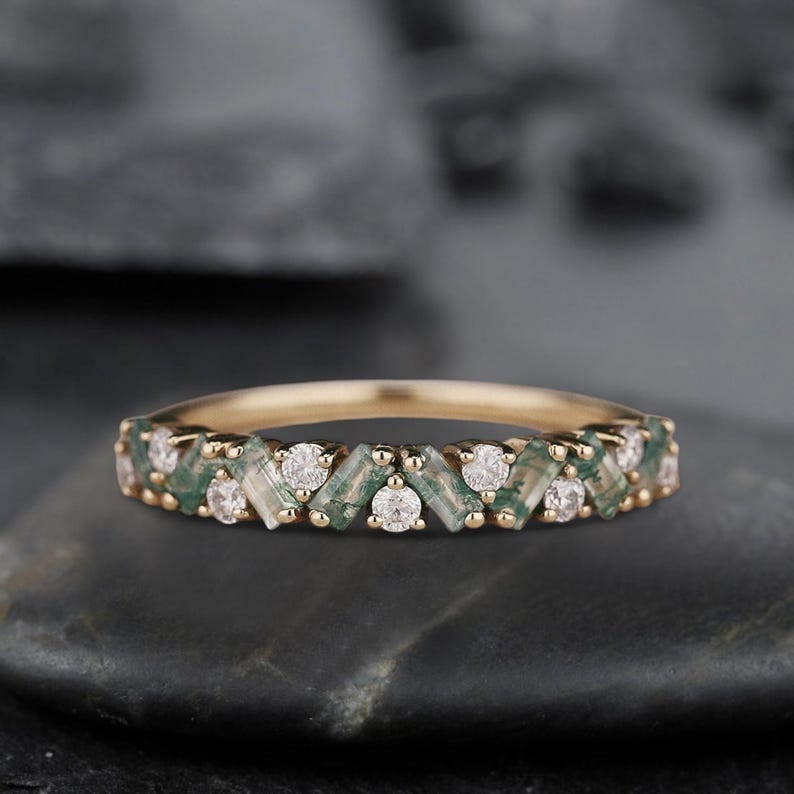 May include: A gold band ring with a row of alternating green and white gemstones. The ring is set on a gray background.