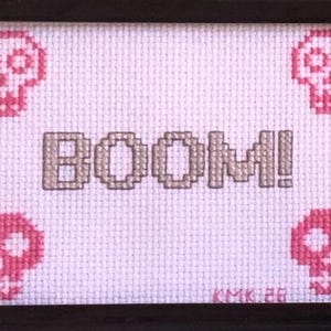 May include: Framed cross-stitch artwork with the word "BOOM!" stitched in tan thread. Four pink skulls are in the corners. The background is white. The artist's initials and year are in the bottom right corner of the framed piece.