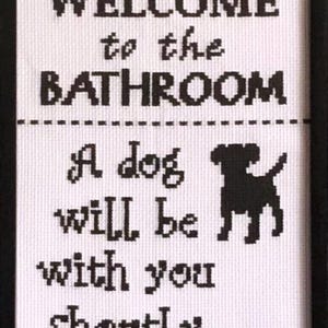 May include: A framed cross-stitch wall art piece featuring black text on a white fabric background. The text reads: WELCOME to the BATHROOM. A dog will be with you shortly... A black silhouette of a dog is included. The piece is in a black frame.