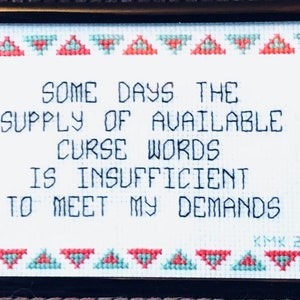 May include: A framed cross-stitch with a white background and black text that reads "Some days the supply of available curse words is insufficient to meet my demands". The text is surrounded by a border of red and green triangles.