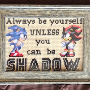 Puede incluir: Un bordado de punto de cruz enmarcado con los personajes de videojuegos Sonic the Hedgehog y Shadow the Hedgehog. El bordado dice "Always be yourself unless you can be SHADOW".