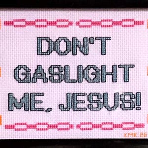 May include: A framed cross-stitch piece on white fabric featuring the text "DON'T GASLIGHT ME, JESUS!" in dark blue block letters. The design is bordered by orange skulls on the left and right sides and horizontal pink chain links at the top and bottom.