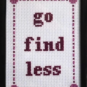 Op de afbeelding: Een ingelijst kruissteekkunstwerk met de woorden "go find less" in donkerpaars op witte stof. Het ontwerp is omrand met paarse kruissteek. De initialen en het jaar van de kunstenaar staan rechtsonder. De lijst is zwart.