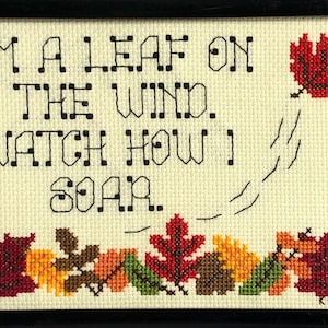 May include: Cross-stitch artwork in a black frame. The piece features the text "I'm a leaf on the wind. Watch how I soar." A red leaf is depicted in the upper right, with a trail of dashed lines. A row of colorful leaves is at the bottom.