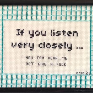 May include: Framed cross-stitch artwork with a black frame. The artwork features the text "If you listen very closely... You can hear me not give a fuck." The background is off-white with a teal and white checkered border.