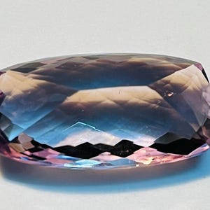 May include: A rectangular-cut gemstone with a range of colours. The gem transitions from a pink-purple at the edges to a deep blue in the centre. The polished surface reflects light, creating a sparkling effect.