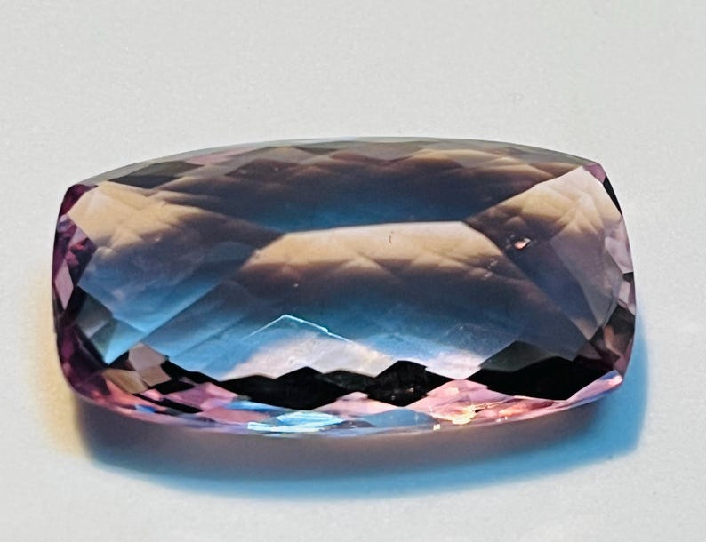 May include: A rectangular-cut gemstone with a range of colours. The gem transitions from a pink-purple at the edges to a deep blue in the centre. The polished surface reflects light, creating a sparkling effect.