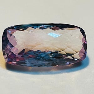 May include: A rectangular gemstone with a faceted surface. The gem transitions from a deep purple to a light lavender hue, with hints of blue. The cut of the stone creates multiple reflective surfaces.