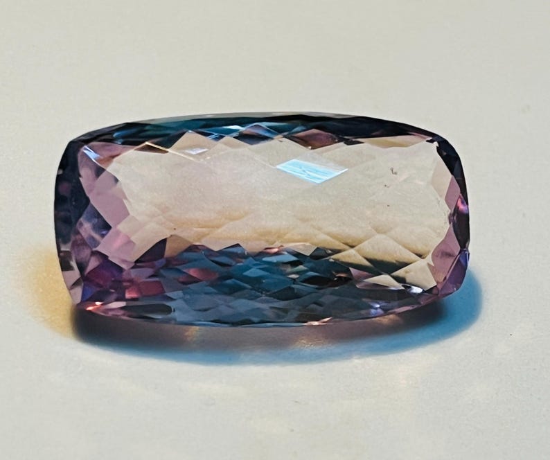 May include: A rectangular gemstone with a faceted surface. The gem transitions from a deep purple to a light lavender hue, with hints of blue. The cut of the stone creates multiple reflective surfaces.