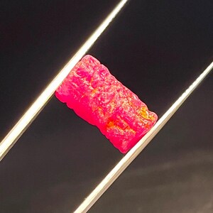 May include: A rectangular, vibrant hot pink gemstone with a rough, textured surface held by a pair of silver-toned metal tweezers. The stone displays deep magenta hues with subtle orange and yellow inclusions across its uneven, crystalline face.