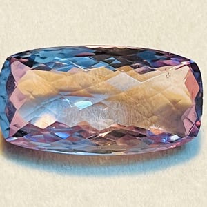 May include: A rectangular-cut gemstone with a colour gradient. The gem's centre is a light, golden-yellow, transitioning to pink and blue at the edges. The cut creates multiple reflective surfaces.