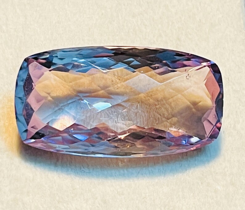 May include: A rectangular-cut gemstone with a colour gradient. The gem's centre is a light, golden-yellow, transitioning to pink and blue at the edges. The cut creates multiple reflective surfaces.
