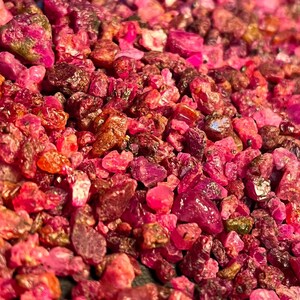 Pode incluir: Uma coleção de pedras preciosas de rubi brutas e não polidas em vários tons de rosa vibrante, vermelho profundo e magenta. As pedras são pequenas, irregulares e angulares, apresentando uma mistura de texturas translúcidas e opacas.
