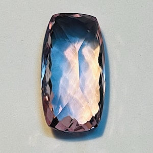May include: A rectangular, faceted gemstone with a gradient of colours. The stone transitions from a deep purple at the edges to a light blue and peach in the centre. The cut of the stone creates multiple reflective surfaces.