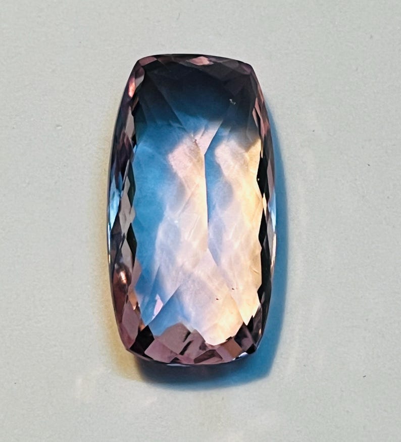 May include: A rectangular, faceted gemstone with a gradient of colours. The stone transitions from a deep purple at the edges to a light blue and peach in the centre. The cut of the stone creates multiple reflective surfaces.