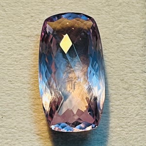 May include: A large, rectangular gemstone with a faceted cut. The gem displays a gradient of colours, shifting from a deep purple at the base to a lighter lavender and blue at the top. A small, yellow diamond-shaped inclusion is visible.