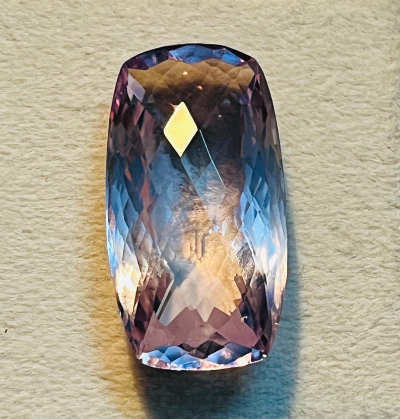 May include: A large, rectangular gemstone with a faceted cut. The gem displays a gradient of colours, shifting from a deep purple at the base to a lighter lavender and blue at the top. A small, yellow diamond-shaped inclusion is visible.
