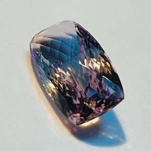 May include: A rectangular-shaped, faceted gemstone. The gem displays a mix of colours, including purple, blue, and pink hues. The facets create a play of light and shadow, enhancing its brilliance. The gem is set against a white background.
