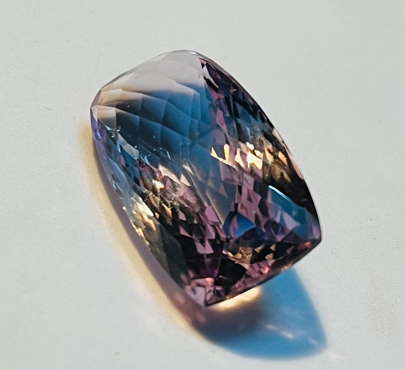 May include: A rectangular-shaped, faceted gemstone. The gem displays a mix of colours, including purple, blue, and pink hues. The facets create a play of light and shadow, enhancing its brilliance. The gem is set against a white background.