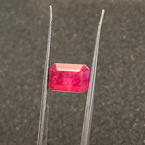 Könnte beinhalten: Ein rechteckig geschliffener Rubin, der von einer Pinzette gehalten wird. Der Rubin zeigt eine tiefe, rote Farbe mit sichtbaren inneren Einschlüssen. Die Facetten des Edelsteins fangen das Licht ein und zeigen seinen Glanz. Die Pinzette ist silberfarben.