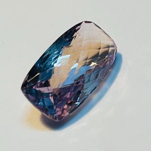 May include: A rectangular-cut gemstone displaying a colour gradient, shifting from deep blue to purple and fading into a pale pink. The facets of the stone reflect light, producing a sparkling effect. This gemstone is suitable for jewellery making.